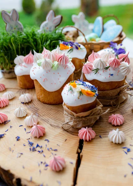 Paskalya dekoru 2021. Beyaz kremalı kekler, pembe bezeli, otlar ve çiçek yapraklarıyla süslenmiş arka planda doğal ahşap arka plan ve yeşillik. Paskalya kartı.