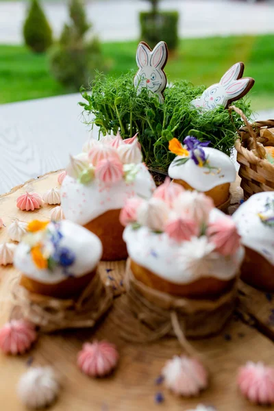 Paskalya dekoru 2021. Beyaz kremalı kekler, pembe bezeli, otlar ve çiçek yapraklarıyla süslenmiş arka planda doğal ahşap arka plan ve yeşillik. Paskalya kartı.