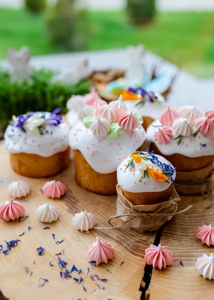 Paskalya dekoru 2021. Beyaz kremalı kekler, pembe bezeli, otlar ve çiçek yapraklarıyla süslenmiş arka planda doğal ahşap arka plan ve yeşillik. Paskalya kartı.