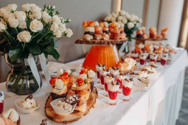 Düğün menüsü. Düğün çikolatası servisi. Tatlı kekler ve kurabiyelerden oluşan bir kompozisyon, fincanlarda tatlılar ve beyaz masa örtüsünde vazoda güller..