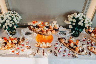 Düğün menüsü. Düğün çikolatası servisi. Tatlı kekler ve kurabiyelerden oluşan bir kompozisyon, fincanlarda tatlılar ve beyaz masa örtüsünde vazoda güller..