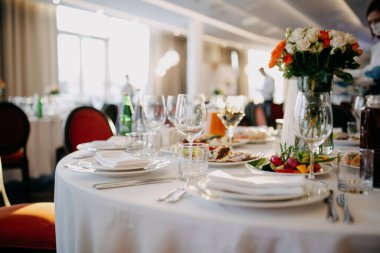 Düğün masasına servis yapıyorum. Restoran işi. Gözlükler, beyaz tabaklar ve yuvarlak bir masada beyaz bir masa örtüsüyle süslenmiş çatal bıçaklar..