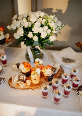 Düğün menüsü. Düğün çikolatası servisi. Tatlı kekler ve kurabiyelerden oluşan bir kompozisyon, fincanlarda tatlılar ve beyaz masa örtüsünde vazoda güller..