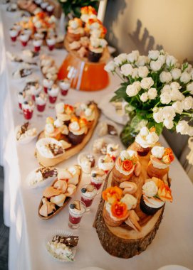 Düğün menüsü. Düğün çikolatası servisi. Tatlı kekler ve kurabiyelerden oluşan bir kompozisyon, fincanlarda tatlılar ve beyaz masa örtüsünde vazoda güller..