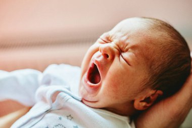 Yenidoğan bebek, yetişkin ellerindeyken geniş ölçüde esniyor, yıldız izli beyaz bir tulum giyiyor, yumuşak bir arka planı var.