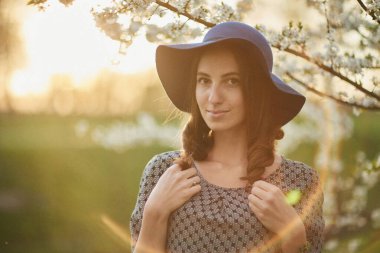 Geniş kenarlı donanma şapkası ve desenli elbisesi olan genç bir kadın altın arkaplan ışıklarıyla çiçeklenen beyaz dallar arasında poz veriyor.