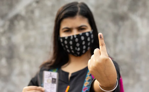 selective focus on ink-marked finger of an Indian woman with safety face mask and voter card on other hand