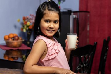 Küçük şirin bir Hintli / Asyalı kızın elinde gülen yüzlü bir bardak süt tutarken fotoğrafı. Sağlıklı beslenme konsepti.