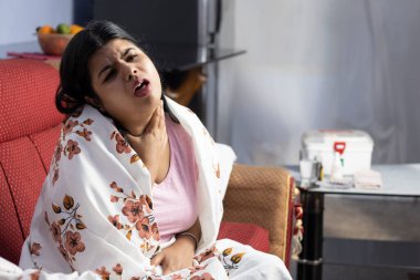 Kanepede oturan, boğazı ağrıyan ve öksüren Hintli bir Asyalı kadın.