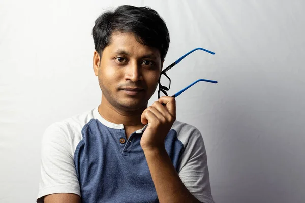 A middle aged Indian man smiling and holding eyeglasses beside his face on white background