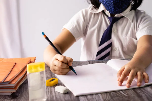 学校の制服と鼻マスクを身に着けているノートに鉛筆で書くインドの子供の手に選択的な焦点 — ストック写真