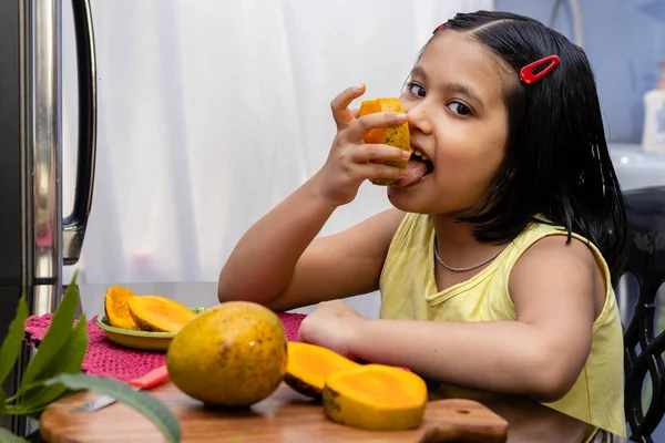 An Indian girl child eating mango and smiling at the camera sitting beside a table; healthy eating concept