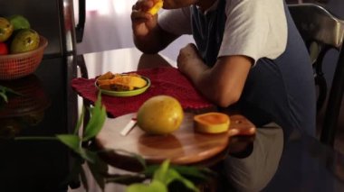 Orta yaşlı, olgun mango yiyen Hintli bir adam bir masanın yanında oturuyor.