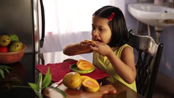 テーブルの横に座ってジェスチャーを示すおいしいマンゴーを食べるインドの女の子 健康的な食事の概念 — ストック動画