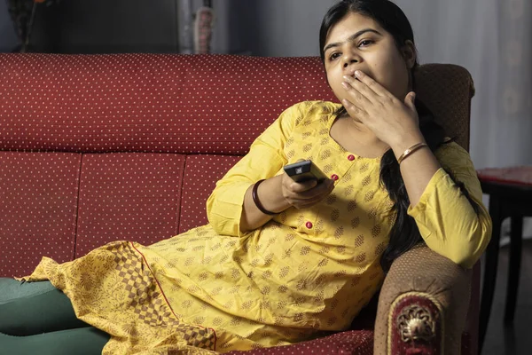 An Indian housewife woman yawning while watching television holding remote in hand, sitting on sofa