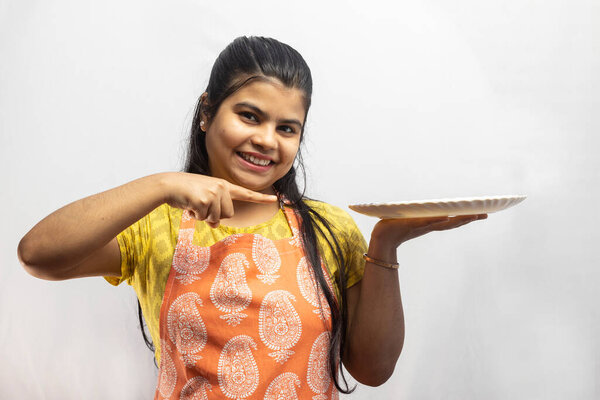 Красивая индийская домохозяйка в фартуке с тарелкой в руке, указывающей на белый фон.
