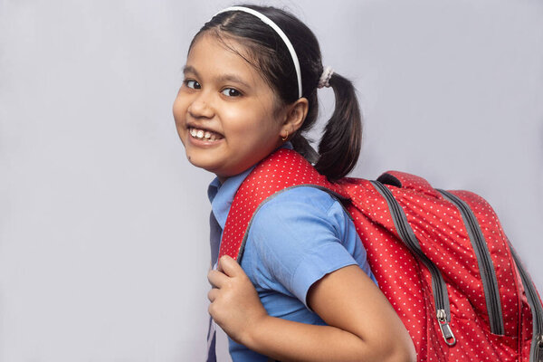 Side view of a happy smiling Indian girl child student in blue school uniform with red bag on grey background