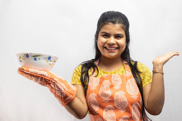 Красивая индийская домохозяйка в фартуке и перчатках с подающей миской в руке улыбается на белом фоне