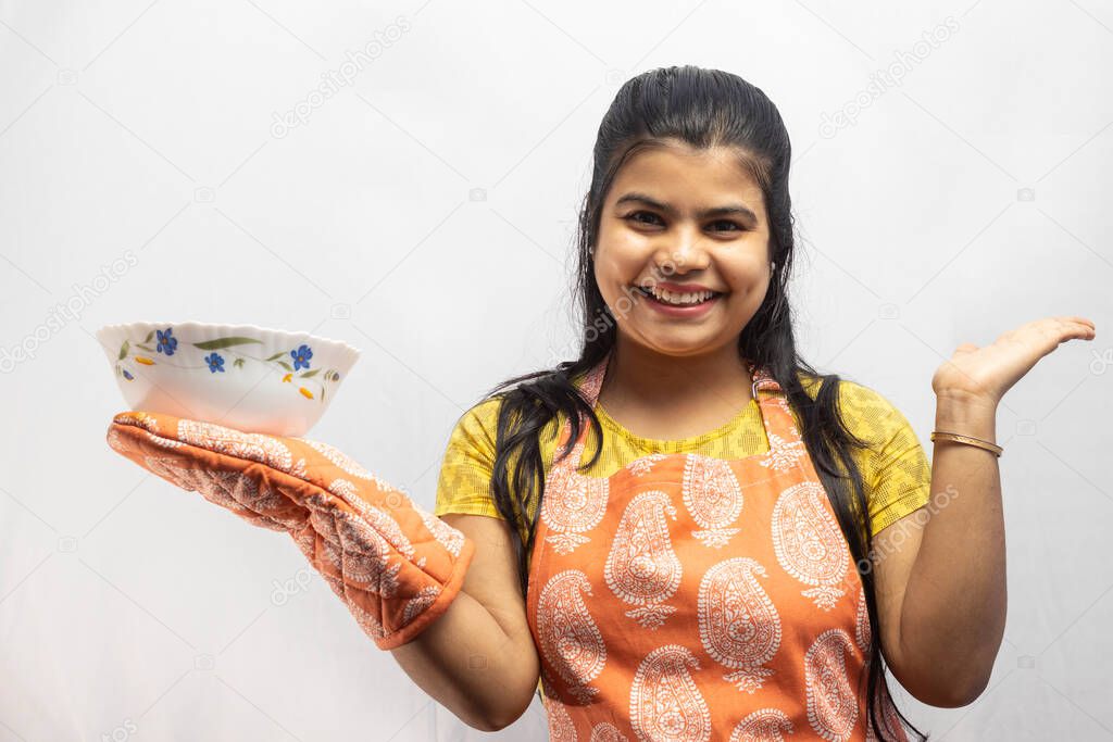 A pretty Indian housewife woman wearing cooking apron and oven gloves with a serving bowl in hand smiles on white background