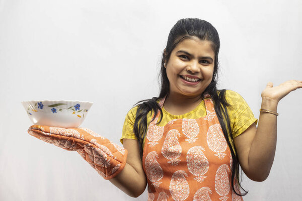 Красивая индийская домохозяйка в фартуке и перчатках с подающей миской в руке улыбается на белом фоне