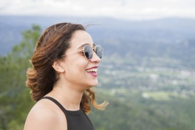 Yan görünüş, güneş gözlüklü, saçları açık genç ve güzel beyaz bir kıza yakın. Gülümseyen ve doğanın tadını çıkaran, ön planda seçici bir odak.