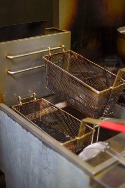 Bir sıra paslı metal kızartma sepetleri kirli bir ticari mutfakta hırpalanmış bir fritözün üzerinde asılı duruyor. Yağlı ve karanlık ortamda ağır gıda hazırlıkları ve endüstriyel mutfak işleri yapılıyor..