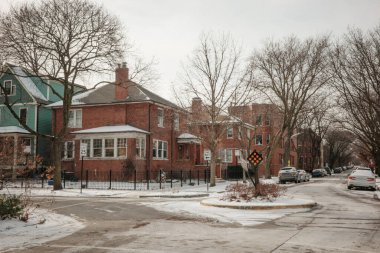 Kış manzarasında kırmızı tuğlalı evler, çıplak ağaçlar ve karla kaplı bir kavşak vardı. Arabalar kaldırım kenarına park edilmiş, sakin, banliyö atmosferi yaratıyorlar..