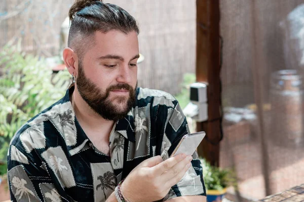 Young man with long hair and beard consults his smartphone in a relaxed way in the garden of his house