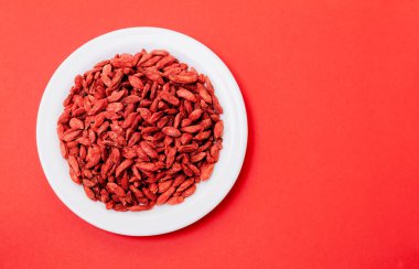 Kırmızı zemin üzerinde beyaz bir tabakta birçok goji kurutulmuş böğürtlen var.