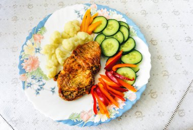 Karnabaharlı domuz boyunlu biftek, yeşil salatalık dilimleri, kırmızı biber şeritleri, masa örtüsündeki bir tabakta.