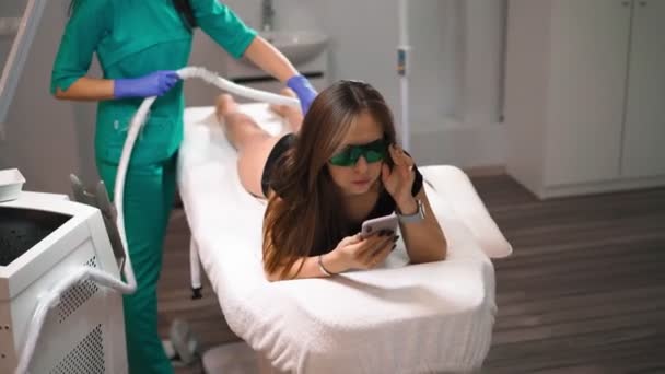 Beautician doet laser ontharing op benen van mooie jonge vrouw die ligt op de medische bank en sms 'en op sociale netwerken via de telefoon in een schoonheidssalon. Cosmitologie en spa-concept — Stockvideo