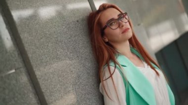 Gözlüklü, uzun kızıl saçlı ve turkuaz takım elbiseli güzel seksi bir kadın şehir merkezindeki bir binanın arka planında duruyor. Açık havada oldukça zayıf bir bayan. Yavaş çekim.