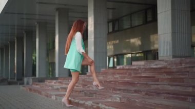 Gözlüklü, kendine güvenen güzel bir iş kadını ve topuklu giysili turkuaz bir takım elbise merdivenleri çıkıyor. Uzun kızıl saçlı seksi bayan girişimci şehir merkezinde ofisin yakınında yürüyor. Yavaş çekim.