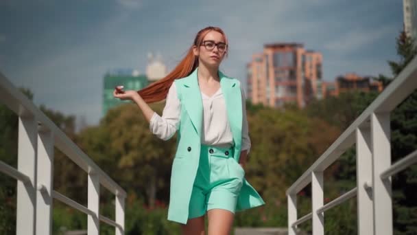戴着眼镜 身穿青绿色时髦西服的漂亮而自信的女商人走着 一位红头发的年轻女企业家走在市中心靠近办公室的地方 慢动作 — 图库视频影像