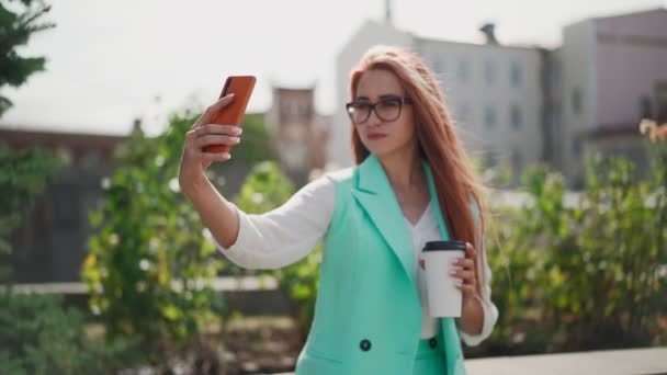 一个戴眼镜的漂亮的现代女人 穿着时髦的蓝绿色西装 手上拿着长长的红头发和咖啡 在城市建筑的背景下 用手机自拍 慢动作 — 图库视频影像