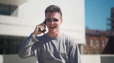 Güneş gözlüklü yakışıklı beyaz adam şehir parkında cep telefonuyla konuşuyor. Dışarıda telefonla konuşurken gülen ve gülümseyen genç adam. Yavaş çekim.