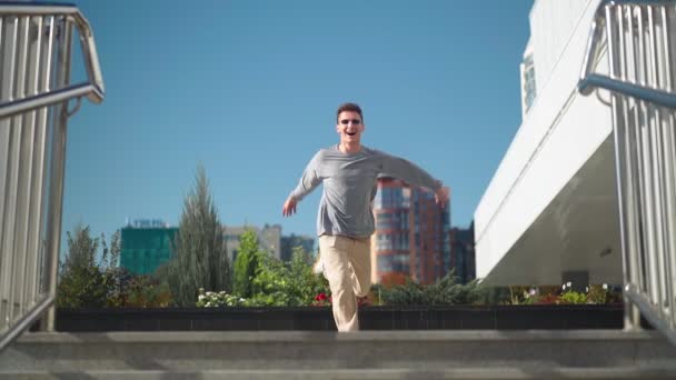 サングラスのハンサムな幸せな面白い男が積極的に実行されている 笑顔と喜びと幸せは 市内の階段を下って行くと踊り ビジネスセンターの隣の都市公園で撮影 スローモーション — ストック動画