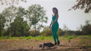 Spor turkuaz tulumlu güzel bir atletik kadın yeşil bir çimenlikte ormanda tüylü kedisiyle yürüyor. Şirin kedi sahibi ile parkta yürüyor. Hayvan bakımı. Yavaş çekim.