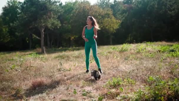 一位穿着绿色运动衫的漂亮的女运动员和她那只毛绒绒的猫在森林的绿茵草坪上散步 可爱的小猫和主人在公园里散步 宠物狗照顾 慢动作 — 图库视频影像