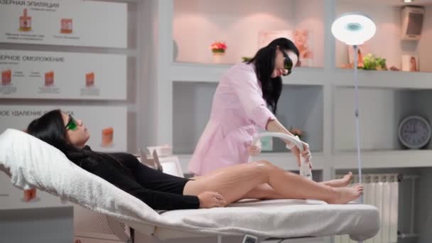 Esteticista femenina haciendo depilación láser en las piernas delgadas de la hermosa mujer sexy que está acostado en el sofá médico en el salón de belleza. Cosmitología, epiléptica y concepto de spa. Cuidado corporal — Vídeos de Stock