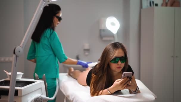 Esthéticienne faisant épilation laser sur les jambes de belle jeune femme qui est allongé sur le canapé médical et textos sur les réseaux sociaux par téléphone dans un salon de beauté. Cosmitologie et concept de spa — Video