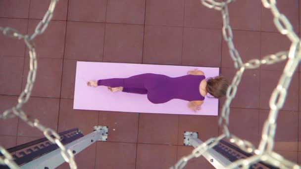 Young athletic woman in a sports purple tracksuit does exercise for stretching while sitting in a split. Fitness outdoors. Slow motion. Top view through a basketball hoop — Stock Video