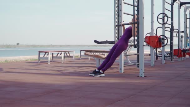 Mujer atlética joven en un chándal deportivo púrpura hace ejercicio pull-up en el bar en el terreno de entrenamiento en un parque de la ciudad. Fitness al aire libre. En cámara lenta. Vista superior — Vídeo de stock