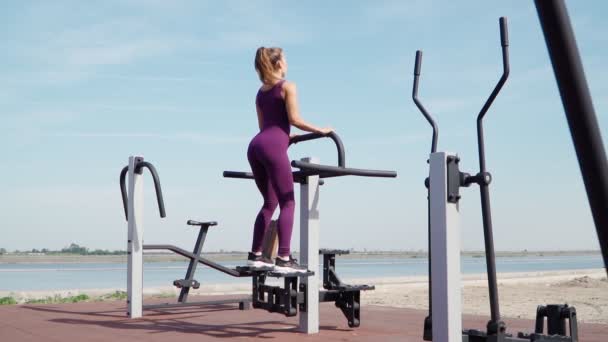 Mujer atlética delgada en un ejercicio de chándal deportivo púrpura en una bicicleta estacionaria en un campo de entrenamiento en un parque de la ciudad. Fitness al aire libre. Movimiento lento — Vídeo de stock