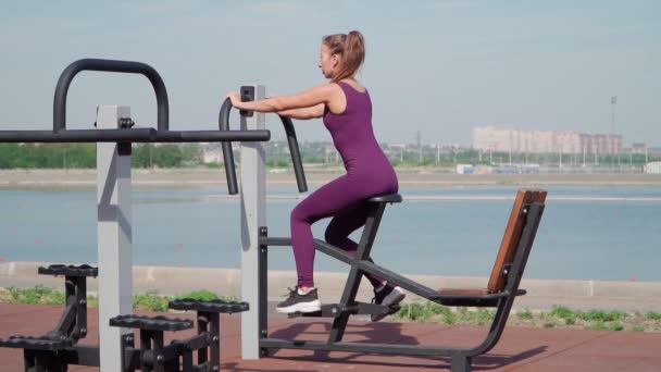 Тонкая атлетичная женщина в спортивном фиолетовом спортивном костюме на стационарном велосипеде на тренировочной площадке в городском парке. Фитнес на открытом воздухе. Медленное движение — стоковое видео