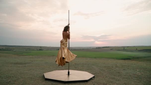 Schöne athletische Frau in einem goldenen Kleid führt Tricks auf der Stange vor dem Hintergrund des Sonnenuntergangs auf dem Feld. Schönheit und Körperpflege. Frauensport und Fitness. Zeitlupe — Stockvideo