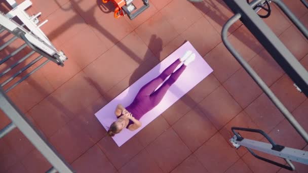 Mujer atlética joven en un chándal deportivo púrpura hace ejercicio de estiramiento en el terreno de entrenamiento en un parque de la ciudad. Fitness al aire libre. En cámara lenta. Vista superior — Vídeo de stock