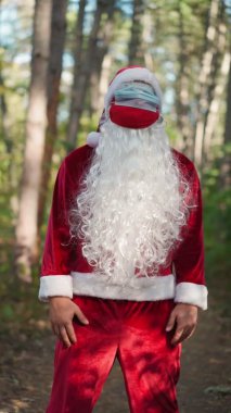 Dikey video. Ormanda yüzünde iki tıbbi maske olan Noel Baba kostümü giymiş komik bir adam. Noel ve koronavirüs. Covid-19. Tatillerde karantina. Yavaş çekim