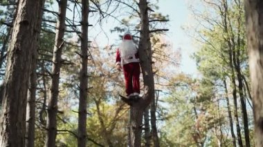 Yüzünde tıbbi maskeyle Noel Baba kostümü giymiş komik bir adam ormanda ağaca tırmandı ve durdu. Noel ve Coronavirus Covid-19. Tatillerde karantina. Yavaş çekim