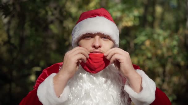 A guy dressed in a suit of Santa Claus puts on red protective medical mask on face in the forest. Coronavirus and Christmas. New Year during quarantine. Slow motion — Stock Video